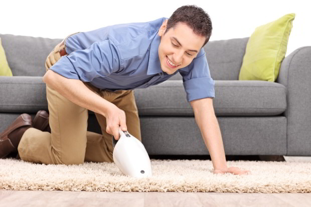 Young joyful man vacuuming a carpet with a handheld vacuum cleaner