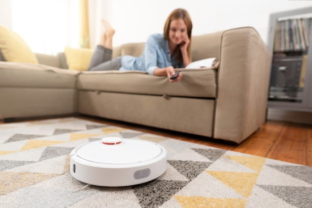Robotic vacuum cleaner cleaning the room while woman relaxing on sofa